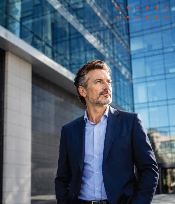 Professional businessman standing outside a modern glass office building, looking confidently into the distance in an urban corporate setting